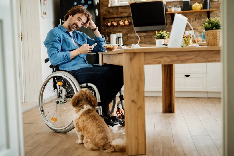 Disabled man in wheelchair sitting at table at home looking at phone and smiling with little dog
